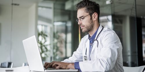 Confident male doctor using laptop at desk in clinic. Medical professional is wearing lab coat. He is working on computer.